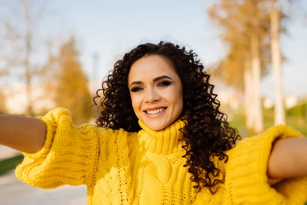 Sarı sonbahar ağaçları ve sarı parlak süveter küçük kıvırcık saçlı kızın üzerinde gülümseyen ve kollarını ileri doğru uzatan.