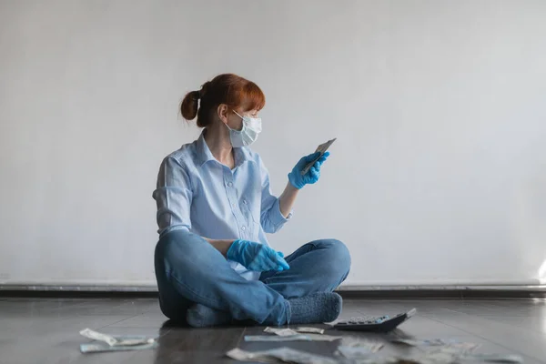 A woman in a medical mask and gloves sits on the floor on a white background and counts money