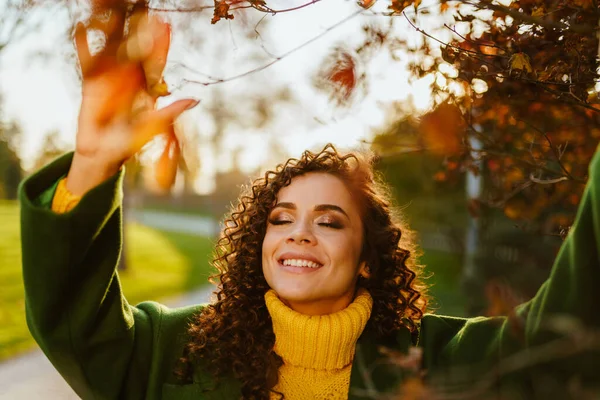 Cubriendo sus ojos con placer y sonriendo con una morena de dientes blancos incluso sonrisa rizada toca las hojas en las ramas del árbol de otoño — Foto de Stock