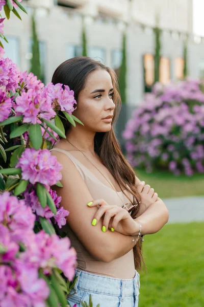 Uma menina bonita com cabelos longos escuros em uma camiseta bege fica nos arbustos floridos no parque. Retrato de uma rapariga de perfil no parque. Summer Park. As meninas olham para a distância. — Fotografia de Stock