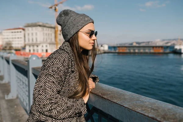 Ritratto di profilo di una ragazza sul ponte sull'argine. Studente con cappello e cappotto grigio, in occhiali neri. Una bella ragazza esamina la superficie dell'acqua durante una passeggiata in Turchia. Giornata di sole. — Foto Stock