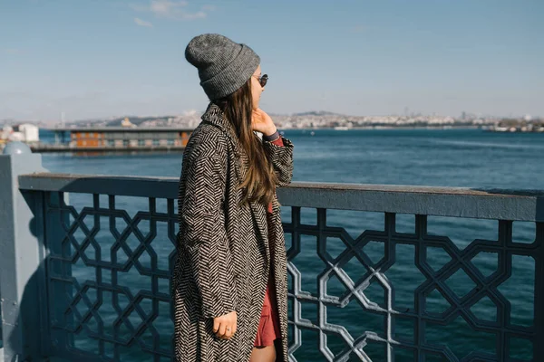 Una ragazza con i capelli lunghi scuri in occhiali, con i cappotti grigi e un cappello guarda il lungomare di Istanbul. Uno studente viaggia durante una pandemia. Passeggiate in autunno all'aria aperta. Foto dal retro, il — Foto Stock