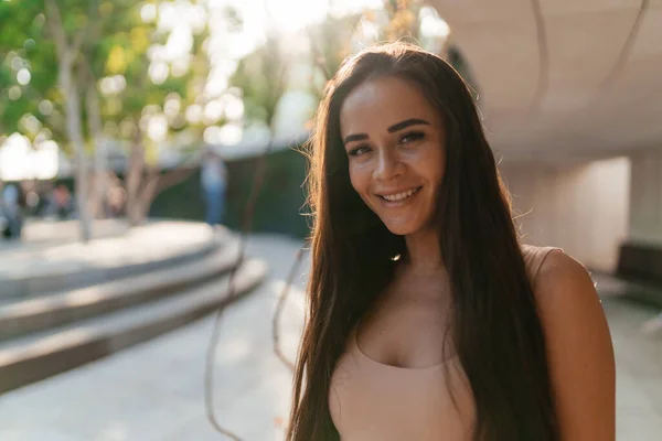 Una joven sonriente con el pelo largo oscuro en una camiseta beige. Retrato de una linda chica de cerca en el pecho. Un disparo contra el sol en un parque verde. — Foto de Stock