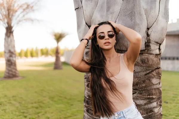 A girl with dark long hair in glasses and a beige T-shirt stands against the background of a palm tree in the park. Walk in the fresh air in the park. Portrait of a girl close-up to the waist. The — Stock Photo, Image