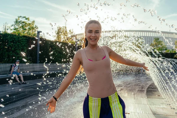 Szczęśliwa dziewczyna w beżowej koszulce i dresach na tle fontanny w parku. Strzał w słońce. Młoda dziewczyna rozłożyła ręce na boki i cieszy się letnim spacerkiem w świeżym — Zdjęcie stockowe
