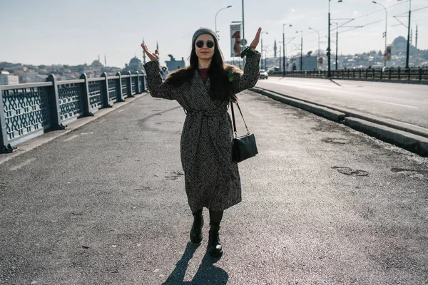 Une jeune fille en manteau gris, chapeau et lunettes noires, leva les mains en marchant sur le pont. Randonnées en Europe. Voyage au début du printemps en Turquie. Météo ensoleillée. — Photo