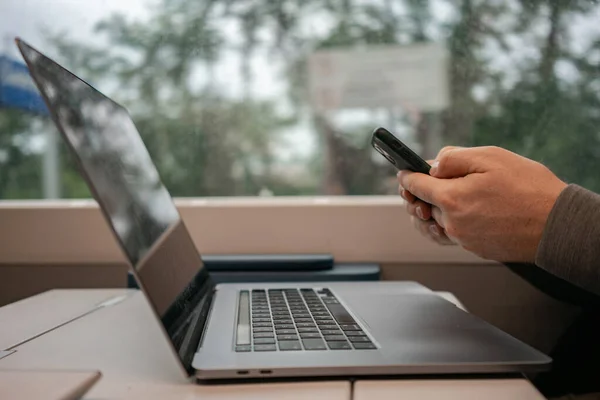 Close-up of a laptop and mens hands typing a message into a phone against the background of a train window. European travel, work on the Internet — Stock Photo, Image