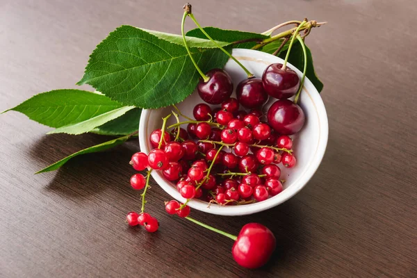 Green cherry leaves, red currant sprigs and ripe cherries in a white bowl closeup. Sweet currants spill out of the bowl. Red cherries and currants. Organic berries - healthy eating and food concept