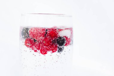 Carbonated drinks with fresh berries and ice. Fresh cold sparkling bubble water with raspberry, black and red currant berries in transparent glass with ice, angle view macro. Food and drink concept