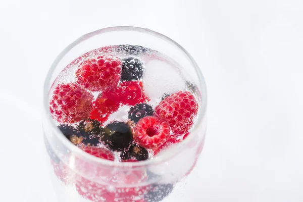 Carbonated drinks with fresh berries and ice. Fresh cold sparkling bubble water with raspberry, black and red currant berries in transparent glass with ice, angle view macro. Food and drink concept
