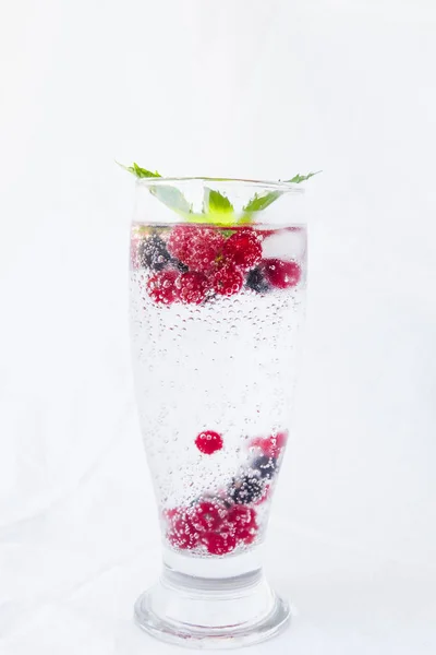 Carbonated drinks with fresh berries, mint and ice. Fresh cold sparkling bubble water with raspberry, black and red currant in transparent glass with ice, angle view, vertical. Food and drink concept