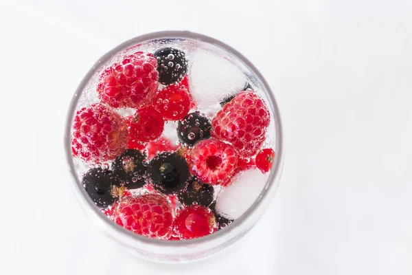 Carbonated drinks with fresh berries and ice. Fresh cold sparkling bubble water with raspberry, black and red currant berries in transparent glass with ice, top view macro. Food and drink concept