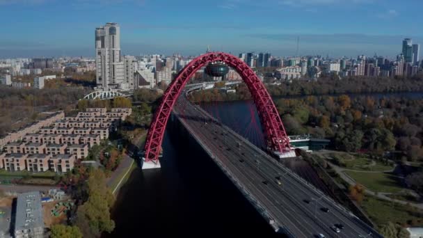 モスクワ 2021年10月1日 川の上の道路上の自動車 駐車場 背後にある都市の建物と現代の都市の赤いケーブルアーチ橋の空中映像 風景橋 美しい街並み — ストック動画