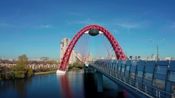 モスクワ 2021年10月1日 川の上の車と自動車のオーバーヘッド道路上のガラスカプセルと近代的な都市の赤いケーブルアーチ橋 都市建築上の空中映像 橋の美術 — ストック動画