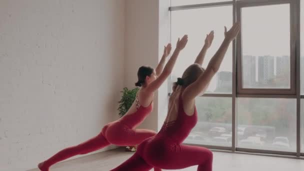 Duas mulheres de uniformes esportivos vermelhos fazem virabhadrasana no estúdio de ioga. Conceito de estilo de vida saudável — Vídeo de Stock