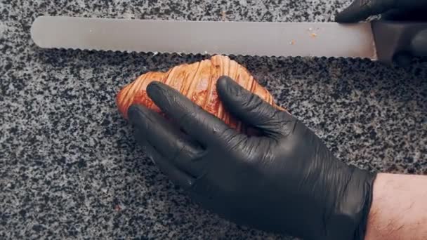 Baker i svarta handskar skär färsk gyllene croissant på mitten på sten bänkskiva. Bageri. Ovanifrån — Stockvideo