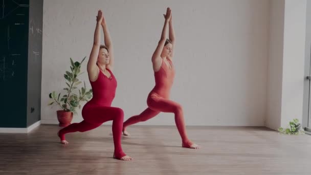 Две женщины в красной спортивной форме делают вирабхадрасану в студии йоги. Понятие здорового образа жизни — стоковое видео