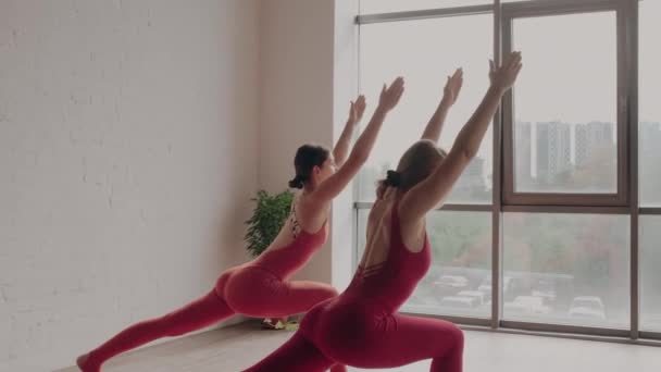 Twee vrouwen in rode sportuniformen maken virabhadrasana in yoga studio. Gezond levensstijl concept — Stockvideo