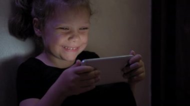 Küçük kız yerde oturuyor ve tablet ya da akıllı telefon oynuyor sosyal internetteki koyu mavi ışıkta blaketin altında.