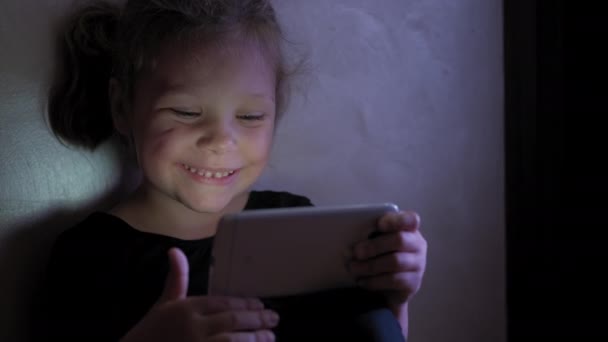 어린 소녀는 바닥에 앉아서 사회적 인터넷에서 어두운 푸른 빛 아래 타블렛이나 스마트폰을 하고 있습니다. 닫아 — 비디오