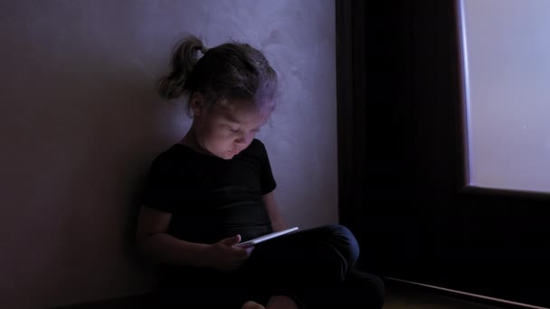 Kleines Mädchen sitzt auf dem Boden und spielt Tablet oder Smartphone im sozialen Internet in dunkelblauem Licht unter schwarzen. — Stockvideo