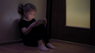 Küçük kız yerde oturup tablet ya da akıllı telefon oynuyor. Sosyal medyada, koyu mavi ışıkta, blaket görüntülerinin altında.