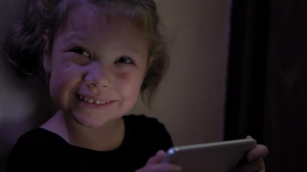 Mała dziewczynka siedząca na podłodze i grająca na tablecie lub smartfonie w internecie społecznościowym w ciemnoniebieskim świetle pod czarnym. Zbliżenie strzału — Wideo stockowe