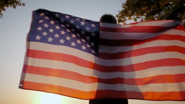 Vista posterior de la mujer feliz con la bandera nacional de EE.UU. de pie al aire libre al atardecer. — Vídeos de Stock