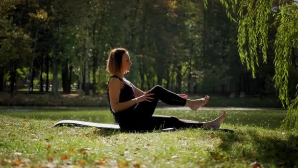 Nő ül jóga szőnyegen, és gyakorolja jóga nyújtás gyakorlat a szabadban a napsütéses napon. Széles látószögű — Stock videók