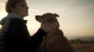 Kapatın. Gün batımında bir köpekle oturan kadın siluetleri.