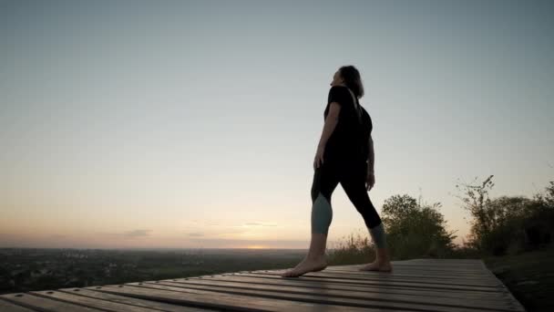 Nő áll jóga szőnyegen és gyakorolja jóga nyújtás gyakorlat a szabadban naplementében. Széleskörű felvétel — Stock videók