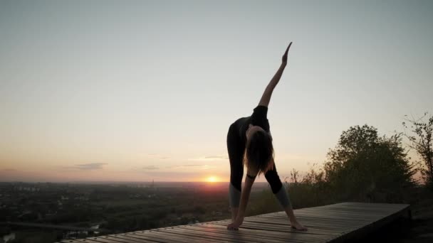 Nő áll jóga szőnyegen és gyakorolja jóga nyújtás gyakorlat a szabadban naplementében. Széleskörű felvétel — Stock videók