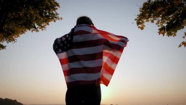 Visão traseira da mulher feliz com a bandeira nacional dos EUA em pé ao ar livre ao pôr do sol. Filmagem em câmara lenta — Vídeo de Stock