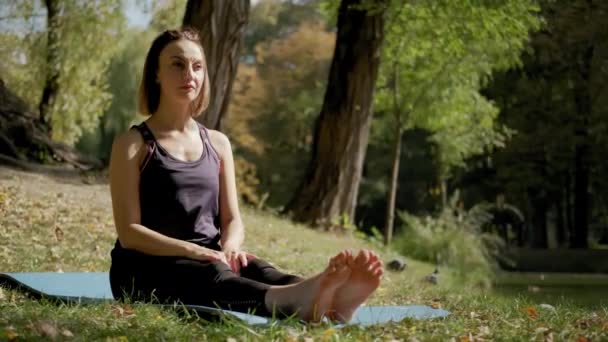 Široký pohled na ženu sedící na podložce jógy a cvičící jógu při cvičení venku za slunečného dne. zpomalený pohyb — Stock video