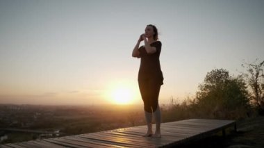 Kadın yoga minderinde duruyor ve gün batımında dışarıda yoga esneme egzersizi yapıyor. Geniş çekim görüntüleri