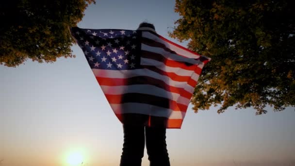 Vista posterior de la mujer feliz con bandera nacional de EE.UU. de pie al aire libre al atardecer. Disparo en cámara lenta — Vídeos de Stock