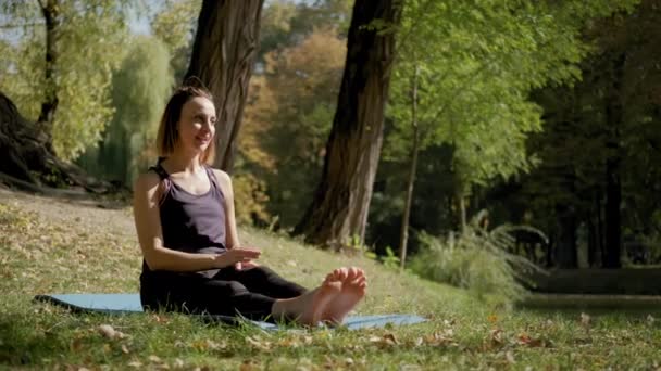Széles kilátás nő ül a jóga szőnyegen, és gyakorló jóga nyújtás gyakorlat a szabadban a napsütéses napon. lassított felvétel — Stock videók