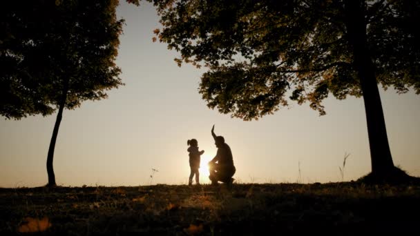 Sobre el fondo del brillante cielo anaranjado del atardecer, la silueta del padre abraza a su hija, la rodea y la arroja en cámara lenta — Vídeo de stock