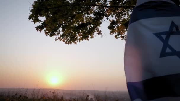 Une femme se tient debout avec le drapeau d'Israël contre un ciel clair au coucher du soleil. Vue rapprochée — Video