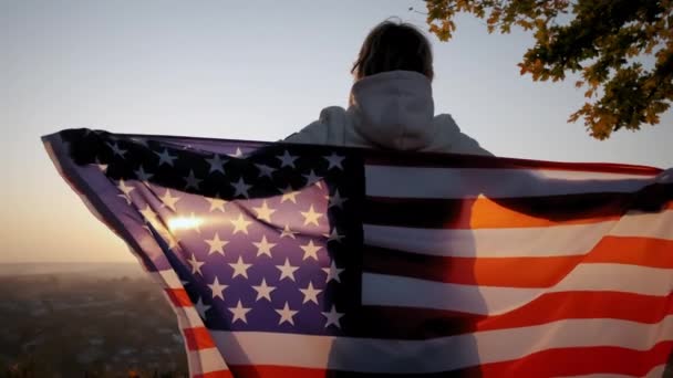 Visão traseira da mulher feliz com a bandeira nacional dos EUA em pé ao ar livre ao pôr do sol. Movimento lento — Vídeo de Stock