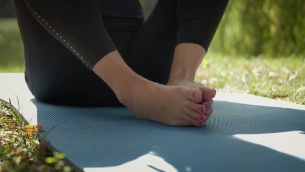 Nő ül jóga szőnyegen, és gyakorolja jóga gyakorlat a szabadban a napsütéses napon. A mezítláb nagyon közel van. — Stock videók