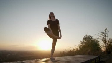 Parlak turuncu günbatımının arka planında gökyüzü kadını yoga minderinde duruyor ve gün batımında dışarıda yoga esneme egzersizi yapıyor.