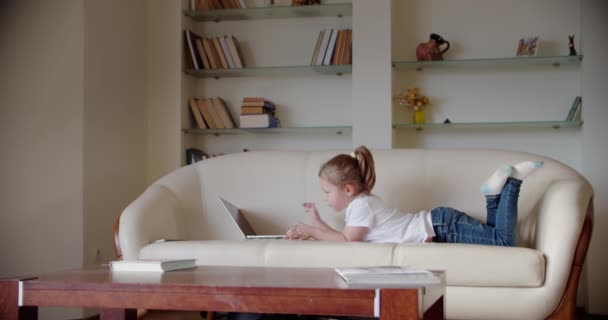 Uma menina fica em casa no sofá e olha para um laptop., joga e assiste o vídeo ou loja na Internet ou escolher produto para encomendar on-line — Vídeo de Stock