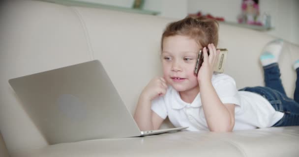 En lille pige ligger hjemme på sofaen og kigger ind i en bærbar computer., og taler i telefon – Stock-video