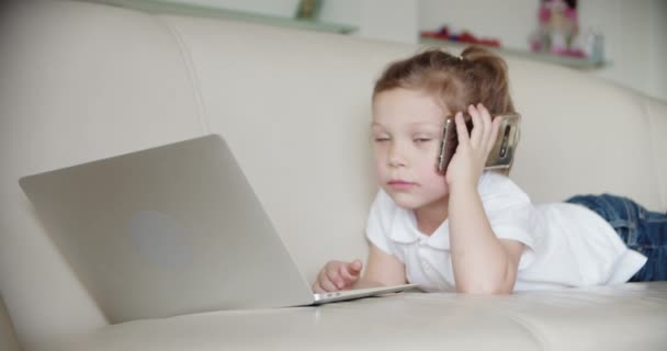 En liten flicka ligger hemma på soffan och tittar in i en laptop., och talar i telefon. Närbild — Stockvideo