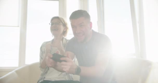 Счастливая пара, сидящая на софе и играющая в видеоигры, с удовольствием играет в онлайн-видеоигры вместе. Медленное движение — стоковое видео