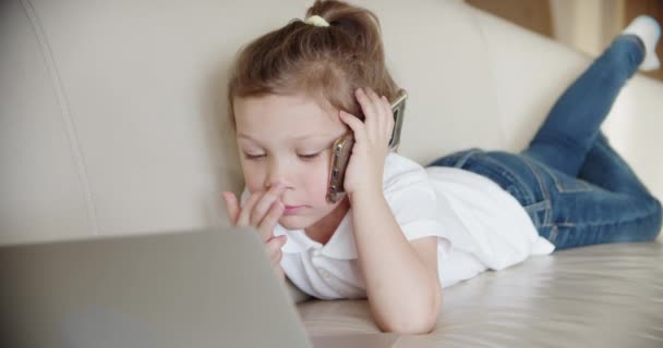 En liten flicka ligger hemma på soffan och tittar in i en laptop., och talar i telefon. Närbild — Stockvideo