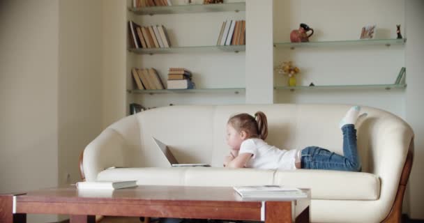 Une petite fille se trouve à la maison sur le canapé et regarde dans un ordinateur portable., joue et regarde la vidéo ou Boutique sur Internet ou Choisir un produit à commander en ligne. Mouvement lent — Video