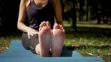 Yoga minderinde oturan ve güneşli bir günde dışarıda yoga egzersizi yapan bir kadın. Çıplak ayaklar çok yakın.