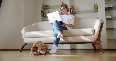 Kadın dizüstü bilgisayar ekranına bak kanepeye otur, kahverengi bir köpek kanepenin yanında yatıyor. Geniş görüntü